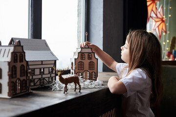 Small girl, standing by windowsill, playing with wooden toy house and deer in dark room with christmas decorations. Winter holidays at home. Leisure activity for children during quarantine time.