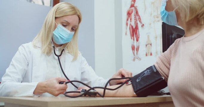 Portrait of Caucasian female healthcare professional in medical mask sitting in clinic and measuring blood pressure of senior patient woman with manometer. Coronavirus infection concept