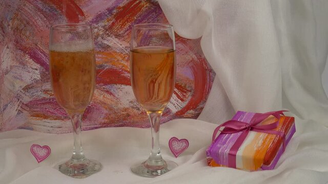 Pink champagne is poured into one of two glasses standing on a white cloth near a gift with lesbian symbols, against the pink and white background 