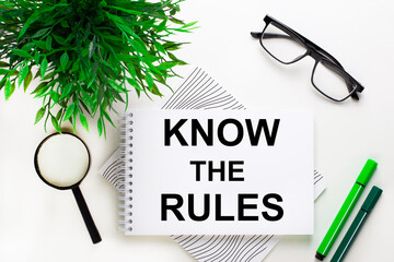 On a white background lies a notebook with the word KNOW THE RULES, glasses, a magnifying glass, green markers and a green plant