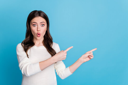 Photo of adorable surprised wavy lady wear white sweater pointing two fingers empty space isolated blue color background