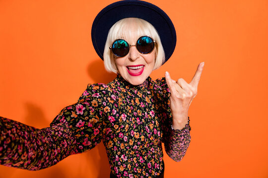 Portrait of old funky crazy smiling good mood lady take selfie showing rock n roll sign isolated on orange color background