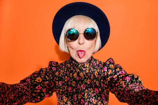 Portrait of old funky cool crazy hipster grandmother take selfie stick tongue out isolated on orange color background