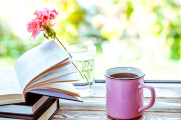 A pink cup with tea stands on a wooden windowsill next to books and a glass in which stands a pink...