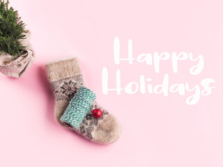 Minimal Christmas flat lay on pink with cozy socks, green twine and wrapped plant tree. Happy holidays greetings text
