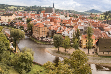 Fototapeta na wymiar Rooftop view to Cesky Krumlov town. Triangle orange roofs and beautiful bell tower. Cloudy sky, green foliage. No people