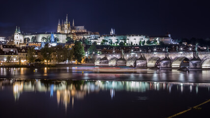Fototapeta na wymiar Night cityscape of Prague, Czech Republic. Stone bridge and embankment, blurred reflections in the river, buildings in the night illumination, a huge cathedral in the background