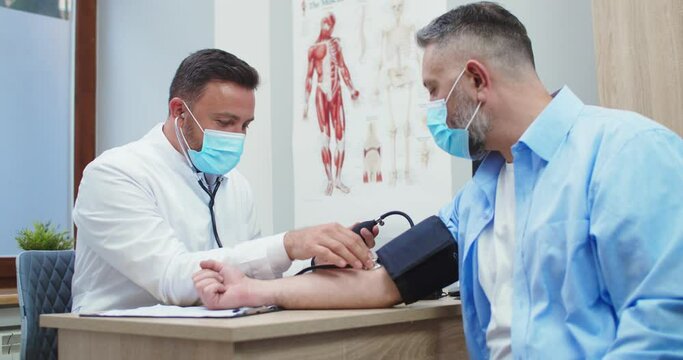 Portrait of Caucasian handsome male doctor in medical mask sitting in clinic and measuring blood pressure and heart rate of middle-aged man patient with manometer. Coronavirus pandemic concept
