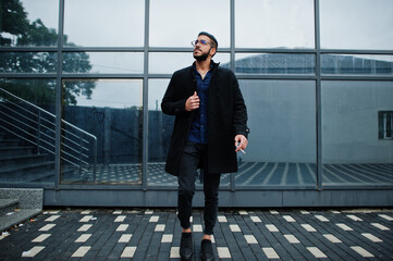 Middle eastern entrepreneur wear black coat and blue shirt, eyeglasses against office building smoking cigarette.