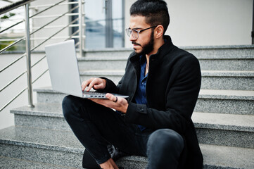 Middle eastern entrepreneur wear black coat and blue shirt, eyeglasses against office building with laptop.