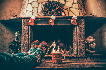 Feet in woollen socks by the Christmas fireplace. Woman relaxes by warm fire with a cup of hot...
