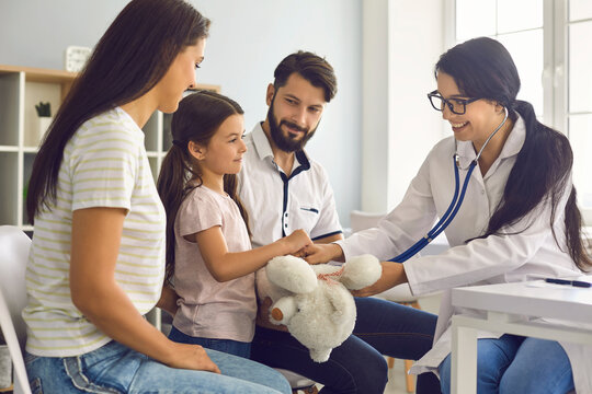 The Family Doctor Listens With A Stethoscope To The Child With The Parents Sitting At The Table In The Clinic.