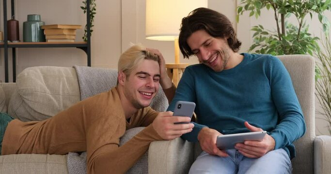 Joyful gay couple using mobile phone and digital tablet at home