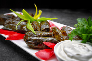 Fototapeta na wymiar Dolma on a white rectangular plate, with herbs, sour cream and bell pepper, on a dark background