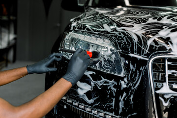 Close up photo of hands of auto service male worker in black protective gloves cleaning car hood and headlights with foam and soft brush. Carwash and detailing. - obrazy, fototapety, plakaty