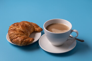 Cup of milk coffee and croissant on blue background, copy space for text
