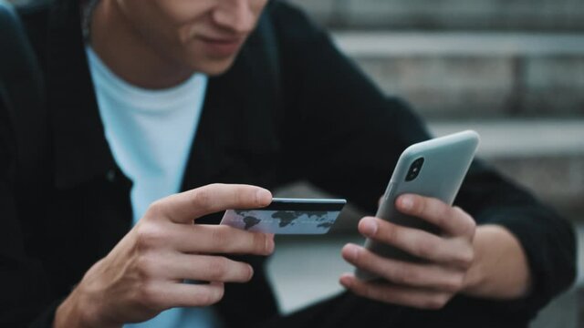 Close up shot of student guy paying online for purchases using smartphone and credit card outdoor