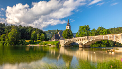 Long exposure sunny nature landscape with mountain lake in Slovenia. beautiful lake Bohinj with perfect sky at summer. view on calm lake with azure water, church and stone bridge on background.