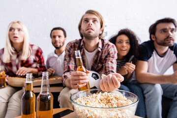 concentrated man taking popcorn while watching football competition near multiethnic friends on blurred background