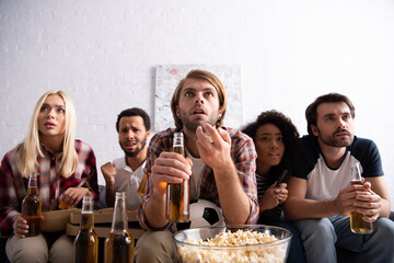 worried man gesturing while holding beer and watching football championship with multiethnic friends