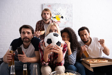 tense woman obscuring face with soccer ball while watching football championship with worried multicultural friends