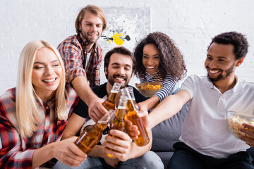 cheerful multicultural friends clinking bottles of beer on blurred foreground