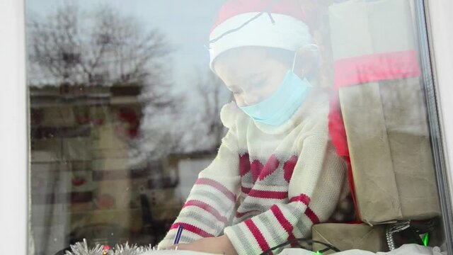 A child in a protective mask writes a letter to Santa Claus on the windowsill. A child in a protective mask writes a New Year's letter during quarantine.A child in a christmas hat looks out the window