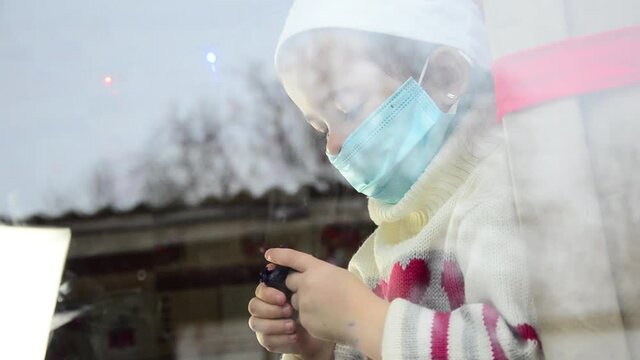 A kid in a mask sits on the windowsill during the New Year holidays. A child wearing a Santa hat looks out the window during New Year's quarantine. Health care concept. Kid plays on the windowsill