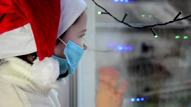 A child wearing a Santa hat looks out a masked window during a Covid-19 quarantine. A small child in a New Year's hat looks out into the street on New Year's holidays. Masked child