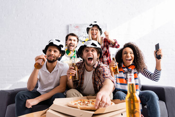 excited man shouting, holding beer and taking pizza while watching championship with multicultural...