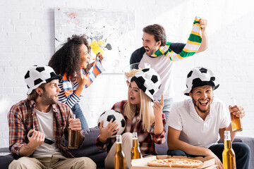 excited woman holding ball and pointing with finger near multicultural friends in football fans hats and scarfs