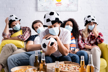 upset man in football fan hat holding soccer ball near sad multiethnic friends in medical masks on blurred background
