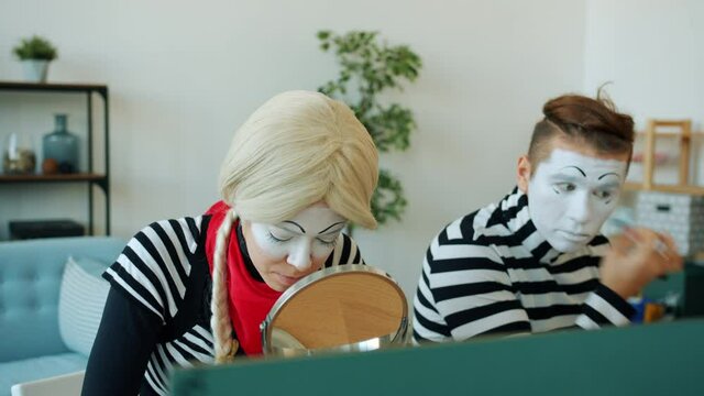 Girl and guy pantomimists are using make-up and face-paint to create performance grimaces indoors in studio. Modern art and creative people concept.