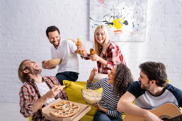 young man playing acoustic guitar to excited multiethnic friends with beer, popcorn and pizza