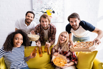 cheerful multicultural friend with beer, pizza and snacks looking at camera during party