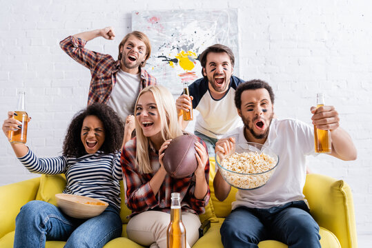 excited multiethnic friends with painted faces holding beer and shouting while watching rugby championship