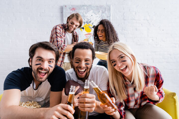 cheerful multicultural rugby fans with painted faces clinking bottles of beer near friends on blurred background