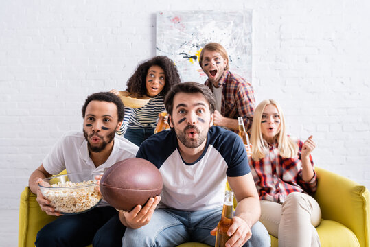 shocked multicultural friends with painted faces watching rugby championship at home
