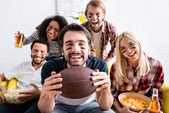 excited man holding rugby ball while watching championship near multiethnic friends with painted faces on blurred background