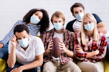 upset man holding rugby ball near sad multiethnic friends in medical masks watching competition at home