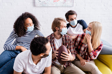 young man holding rugby ball while talking with multicultural friends in medical masks
