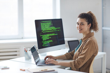 Portrait of young developer smiling at camera while working on computer at her workplace at office