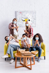 multicultural friends in medical masks chatting on smartphones near table with beer and snacks