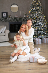 Family - mom, daughter and son, all dressed in white knitted clothes, celebrate the New Year, Christmas. They sit on the floor against the backdrop of New Year's decorations and eat marshmallows.