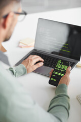 Rear view of man sitting at the table and programming software on laptop and mobile phone