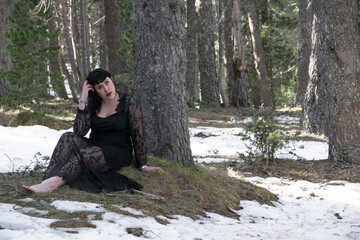 Woman in a dress sitting next to a tree in a snowy forest