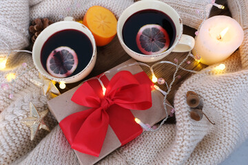 two cups of hot mulled wine with lemon, persimmon and tangerine fruits, candles, box with gift in kraft paper, with red bow lies on table, concept of holiday, congratulations merry christmas, new year
