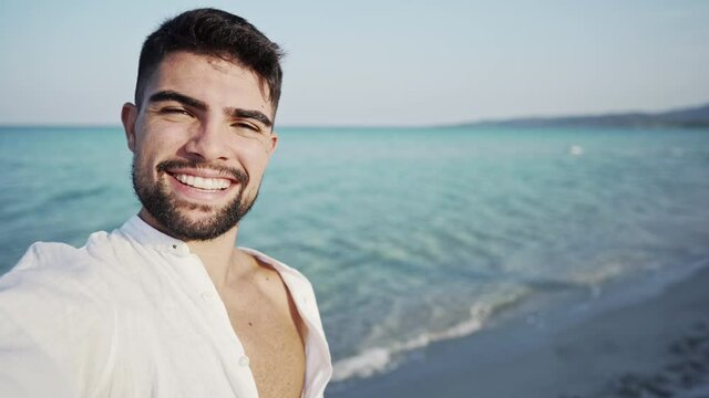 Caucasian handsome bearded guy filming himself in a tropical beach wearing an open white shirt smiling at the sunset - Solo traveler in sea vacation using smartphone to share his happiness for trip