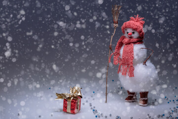 A snowman in a hat and scarf sweeps the snow and looks at the red gifted box. Smooth Black gradient...