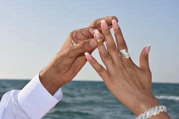 Hands of the newlyweds at the wedding, the groom in a white shirt puts a gold wedding ring on the bride's finger. Blue sea and sky background